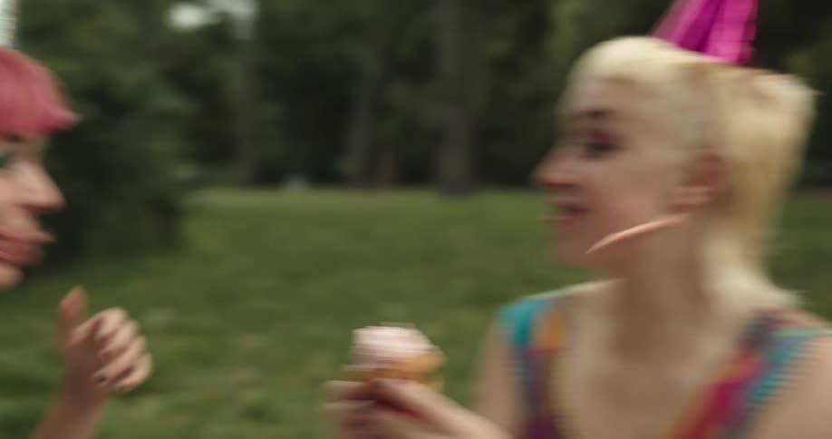 Three women sit together eating cupcakes outside wearing party hats