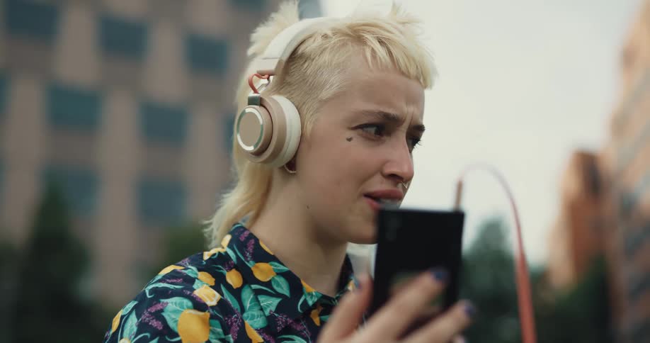 A young woman standing outdoors makes animated facial expressions while using her smartphone for a video chat