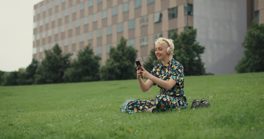 Blonde woman sitting on lawn near building wears headphones while video calling