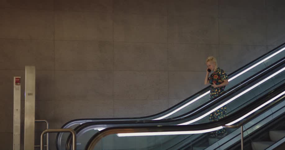 A teenage girl is talking on her cell phone as she rides down an escalator and walks in a hallway