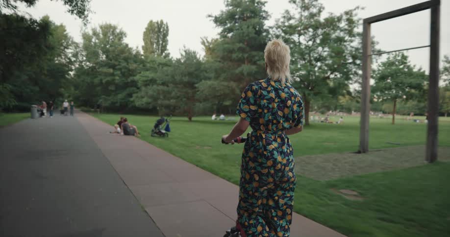 Blonde woman wearing a dress drives an electric scooter through the park at evening