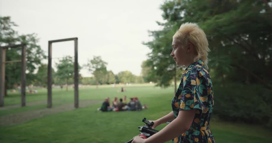 A woman rides an electric scooter through a park as people hang out and picnic