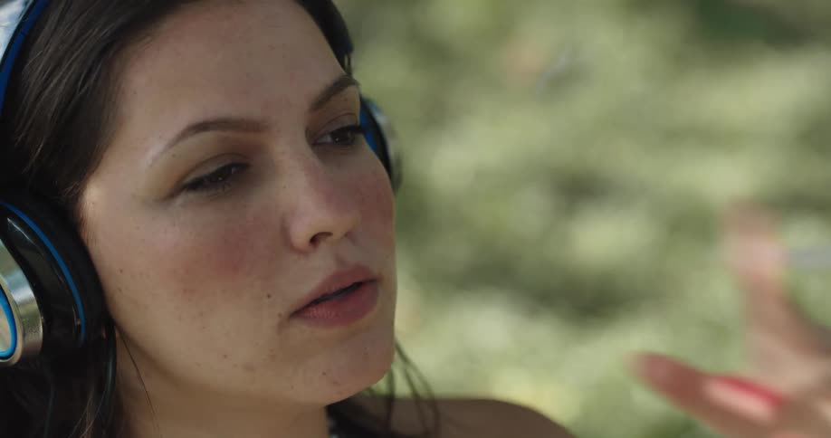 A young woman wearing headphones while sitting against a tree nods her head to the beat while smoking a cigarette