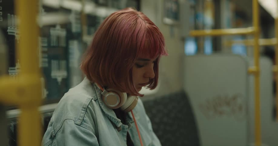 A teenage girl with pink hair and headphones around her neck is sitting on a train reading a book