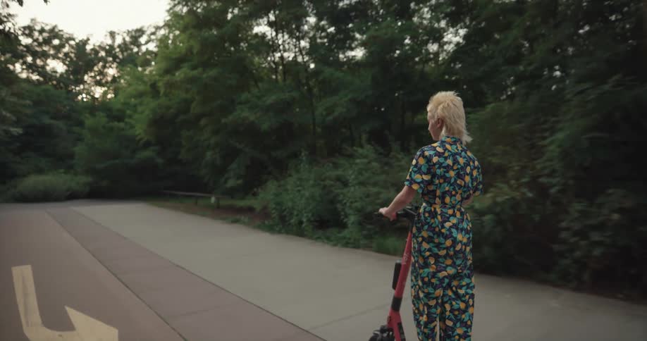 Blonde woman wearing a dress drives an electric scooter through the park at evening