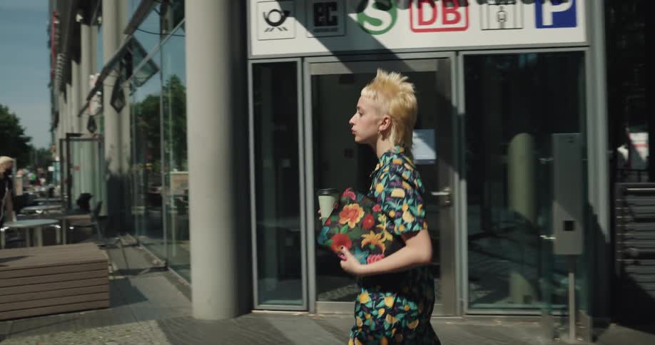 A woman carrying a coffee cup walks down a city street past stores and cafes