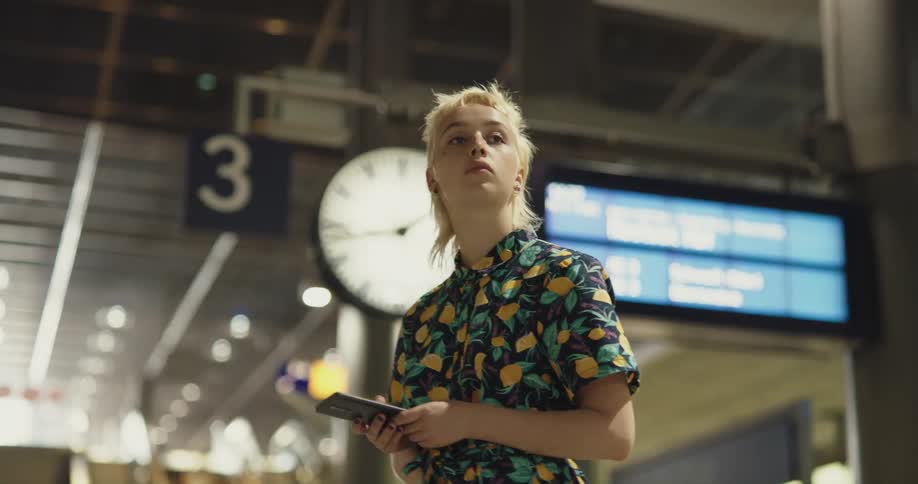 A young woman using a smartphone while standing in a subway station looks up check her surroundings before returning her attention to her phone
