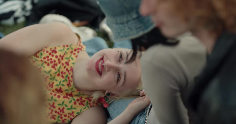 A teenage girl lies on the ground with her head in her friends lap and they talk as her friend strokes her hair