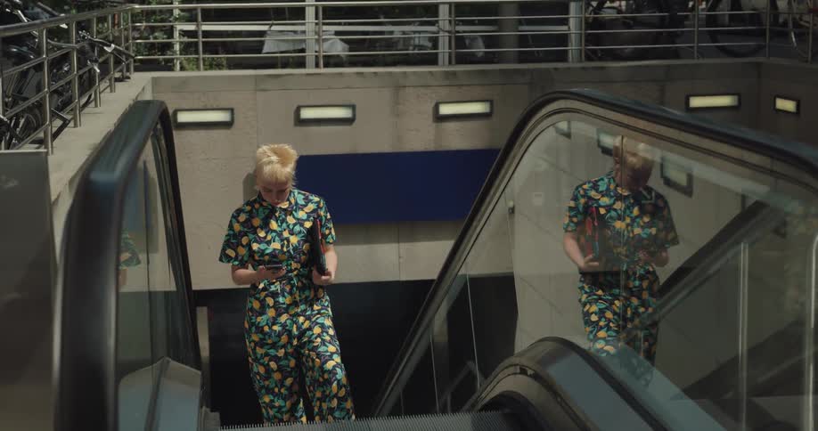 A young woman checks her smartphone while riding an escalator out of a subway during the day