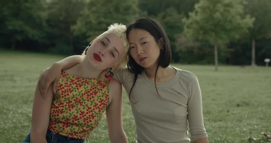 Two girls sit on the grass with their heads resting together while one girl has her arm around the other and then they look in to one anothers eyes and smile