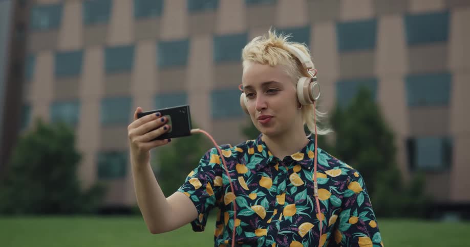 A young woman wearing headphones uses her smartphone to participate in a video chat while sitting in a field outside a building