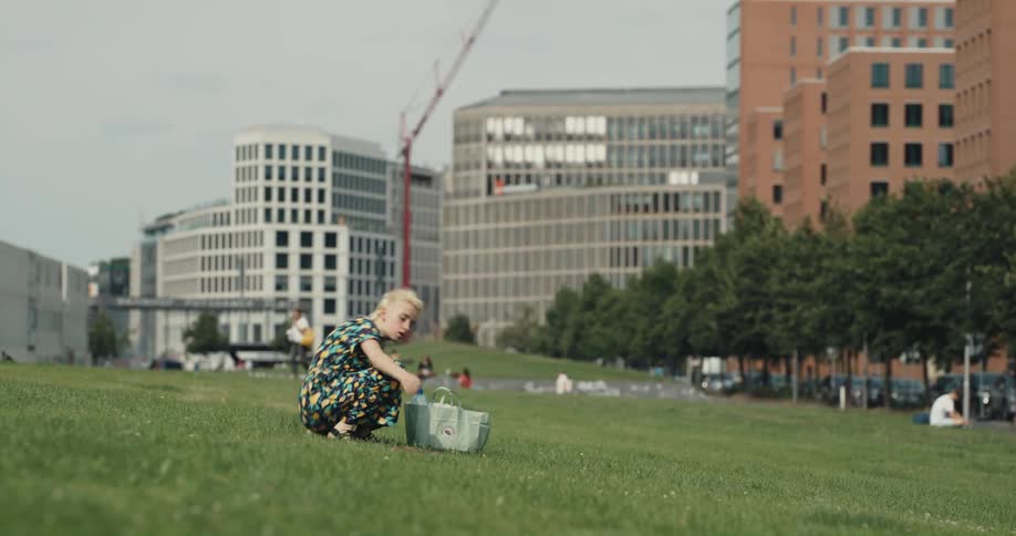 A woman walks away from an apartment building and sits on a grassy hill behind office buildings as other people walk around