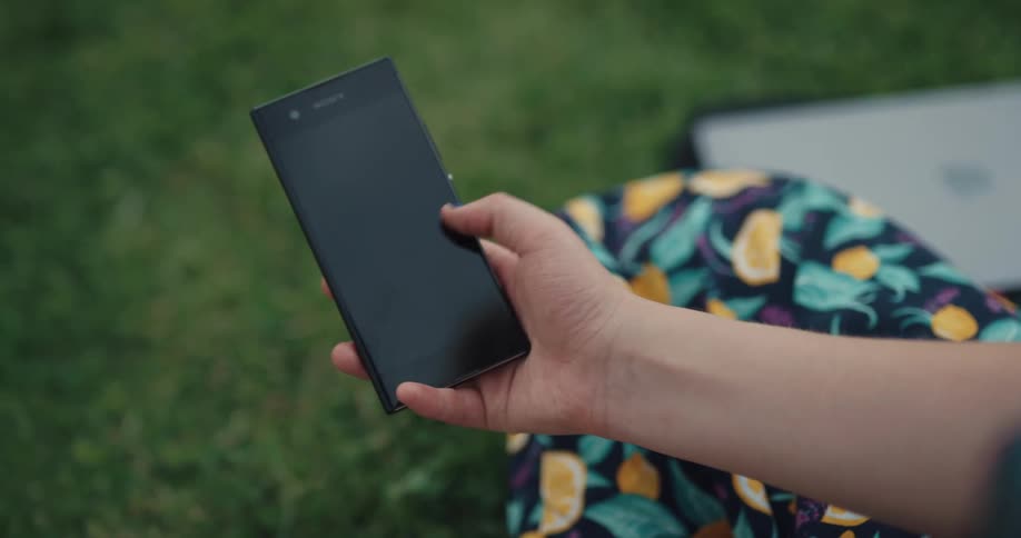 Woman sitting on grass uses holds smartphone in public park
