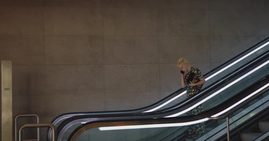 A teenage girl is walking down an escalator and through a hallway as she uses her cell phone