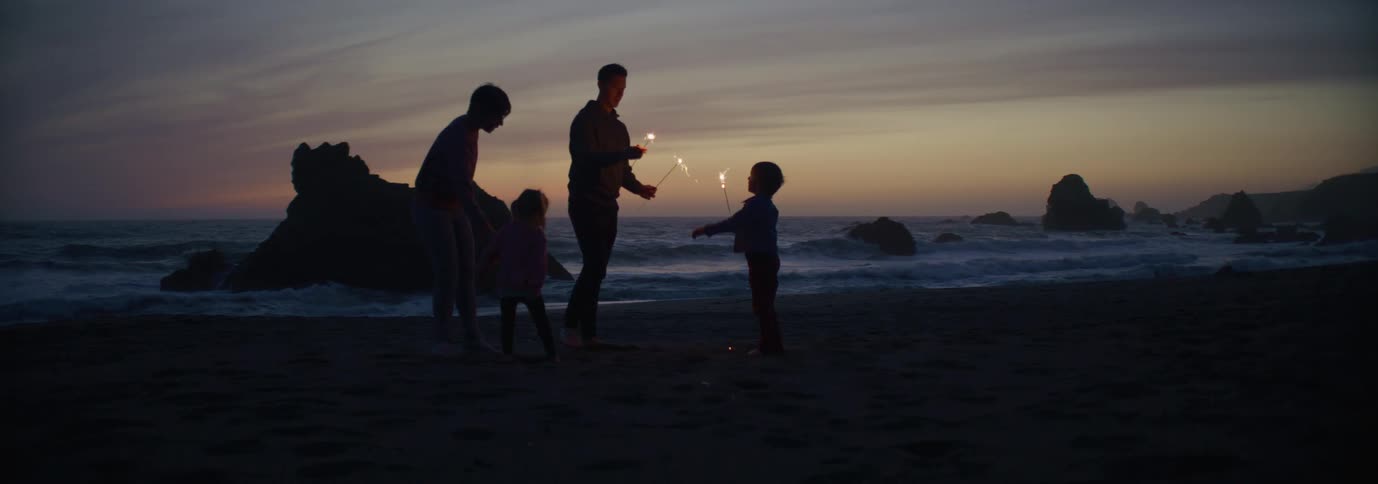 Silhouettes of a family on a beach lighting sparklers for small children as the sun has set
