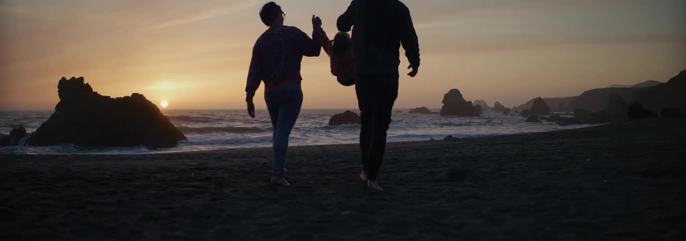 A mother and father hold their little girls hands and swing her as they walk across the beach as waves roll in and the sun sets