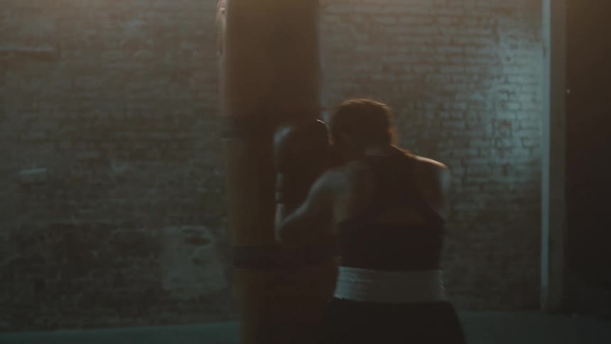 Determined female boxer with braids sports bra fight trunks and boxing gloves hits puching bag in the gym