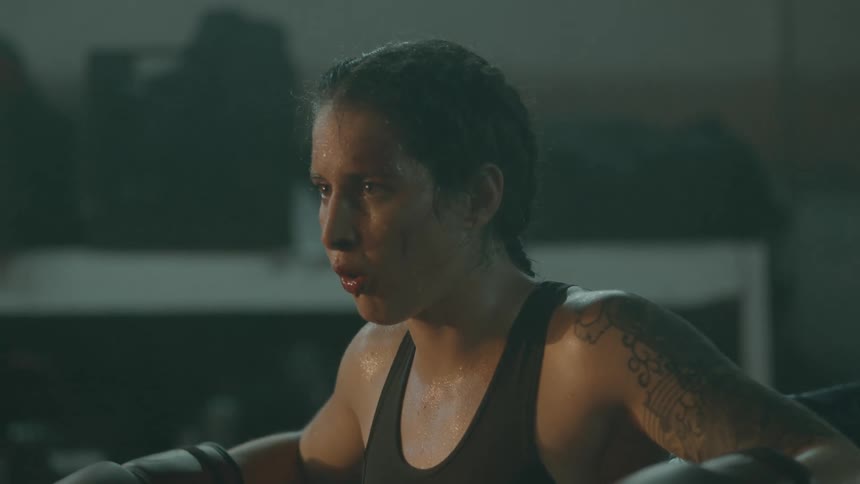Female boxer with braids tattoo sports bra and boxing gloes rests in the corner of boxing ring