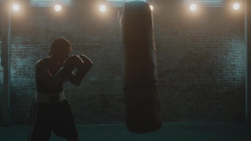 Female boxer with braids sports bra fight trunks and boxing gloves hits puching bag in the gym