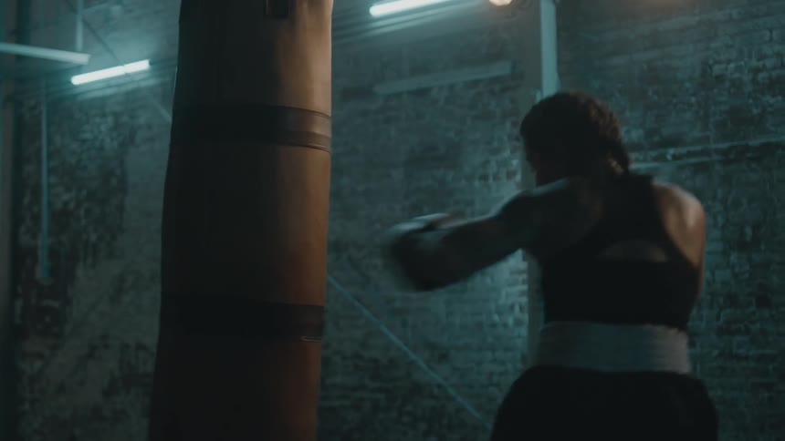 Female boxer with braids sports bra fight trunks and boxing gloves hits puching bag in the gym