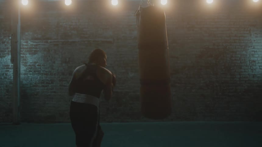 Female boxer trains with body bag in brick walled dimly lit gym