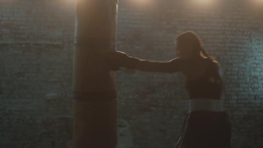 Female boxer with braids sports bra fight trunks and boxing gloves hits puching bag in the gym