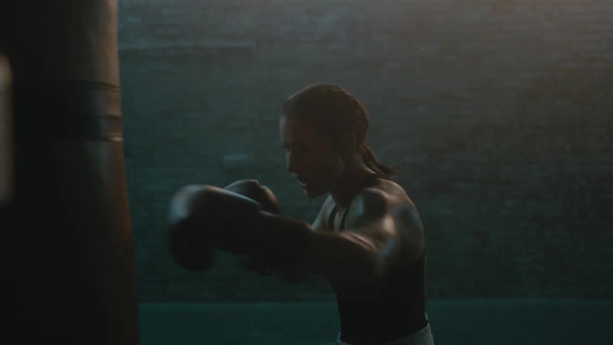 Female boxer with braids sports bra fight trunks and boxing gloves hits puching bag in the gym