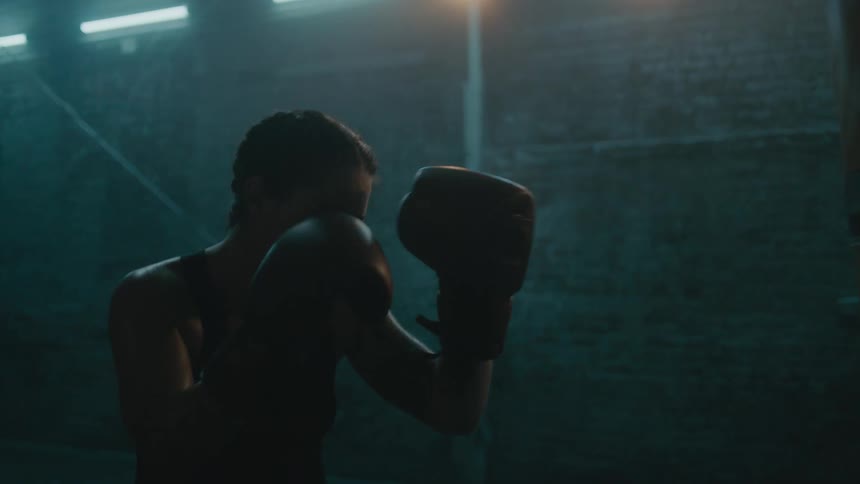 Female boxer sweats while punching heavy bag in dimly lit gym