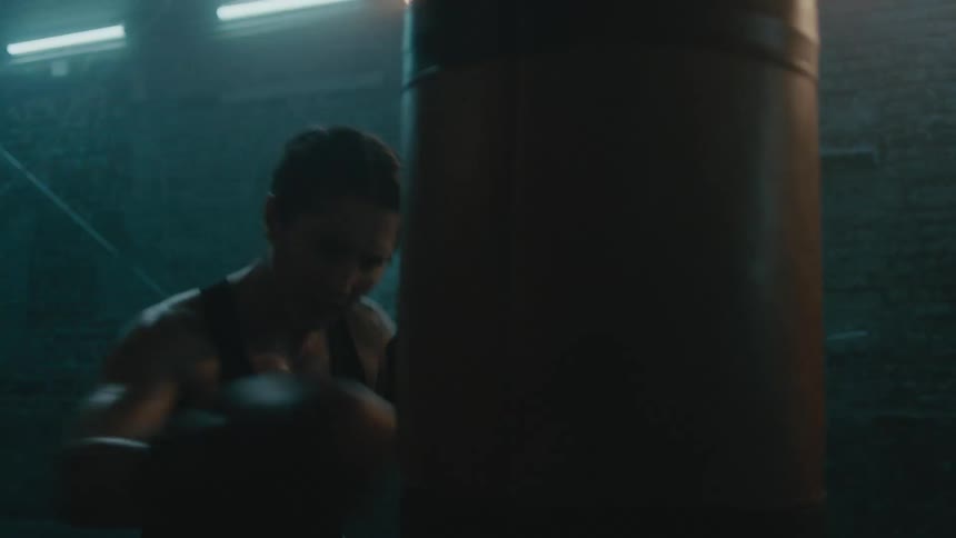 Determined female boxer with braids wearing a sports bra, fight trunks, and boxing gloves hits punching bag in the gym