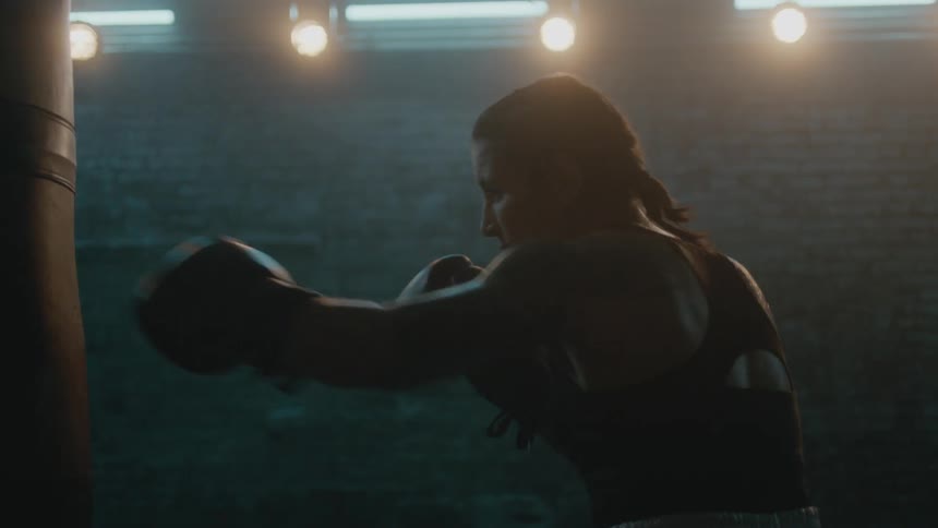 Female boxer with braids wearing a sports bra, fight trunks, and boxing gloves hits punching bag in the gym