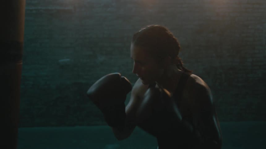 Female boxer with braids wearing a sports bra and fight trunks hits punching bag in the gym