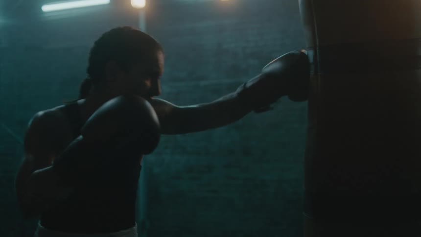 Female boxer with braids wearing a sports bra, fight trunks, and boxing gloves hits punching bag in the gym