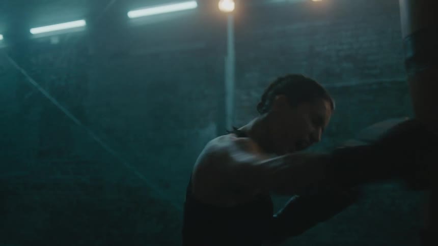 Female boxer with braids wearing a sports bra, fight trunks, and boxing gloves hits punching bag in the gym