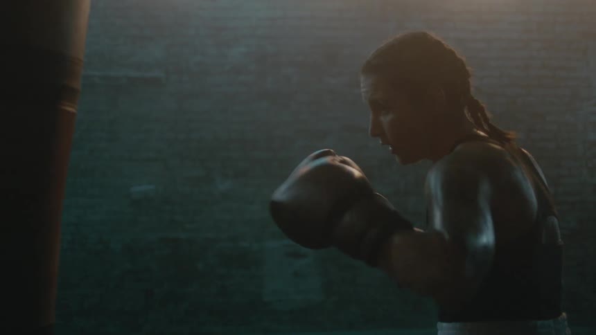 A female boxer repeatedly strikes a punching bag in a dimly lit room