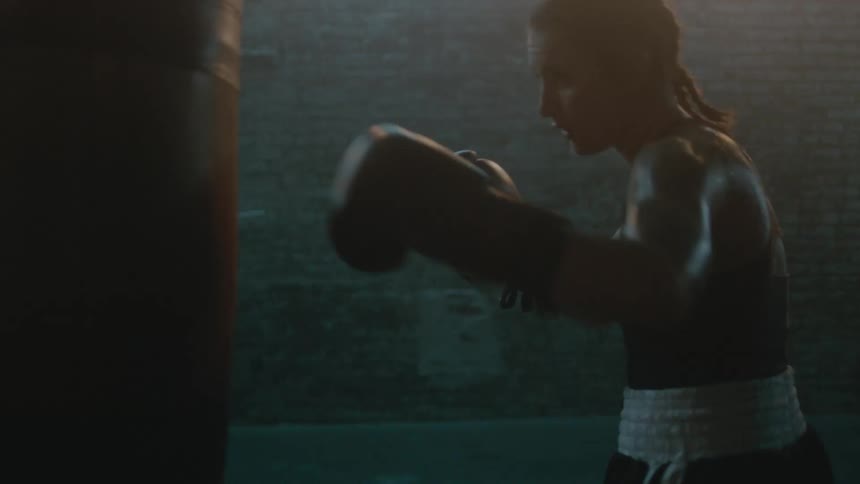 Determined female boxer with braids wearing a sports bra, fight trunks, and boxing gloves hits punching bag in the gym