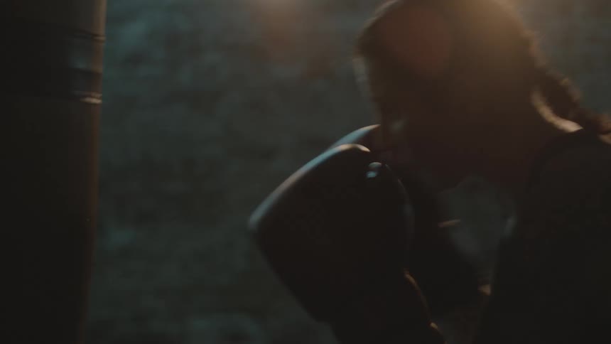 Determined female boxer with braids sports bra fight trunks and boxing gloves hits puching bag in the gym