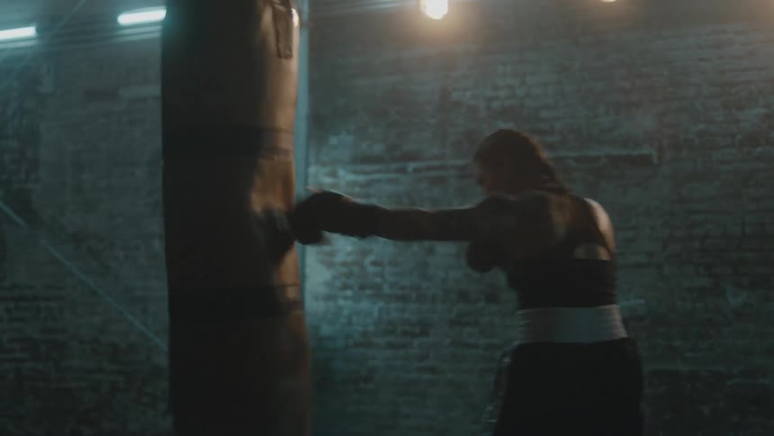 Female boxer with braids sports bra fight trunks and boxing gloves hits puching bag in the gym
