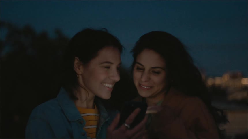 Couple of happy friends talking and laughing looking at a phone in the city at night