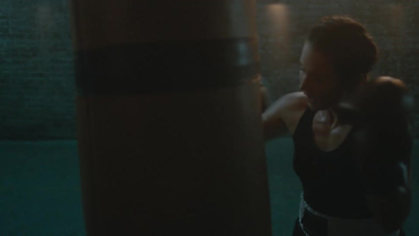 Female boxer with braids wearing a sports bra, fight trunks, and boxing gloves hits punching bag in the gym