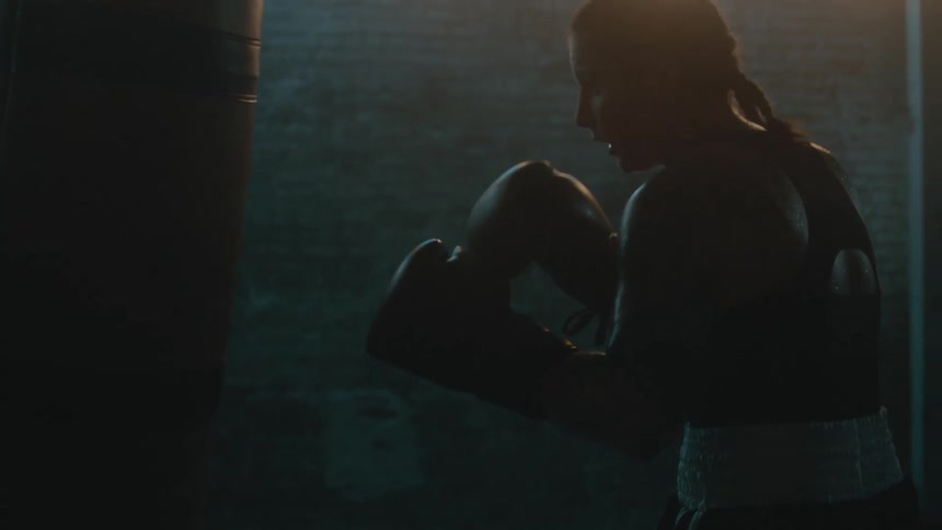 Female boxer with braids wearing a sports bra, fight trunks, and boxing gloves hits punching bag in the gym