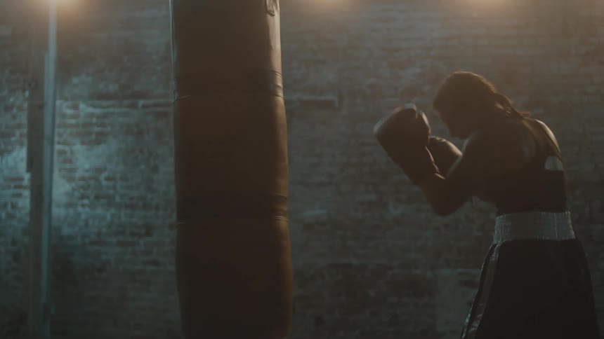 A female boxer practices punches using a punching bag in a dimly lit room