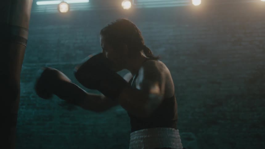 Female boxer with braids wearing a sports bra, fight trunks, and boxing gloves hits punching bag in the gym