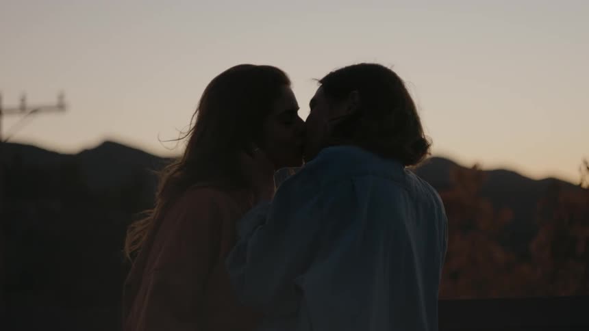 Two women are kissing outdoors as the sun is setting