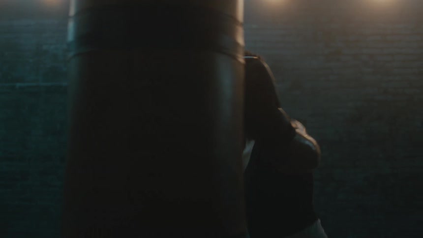 A female boxer circles a punching bag while punching it in dimly lit room