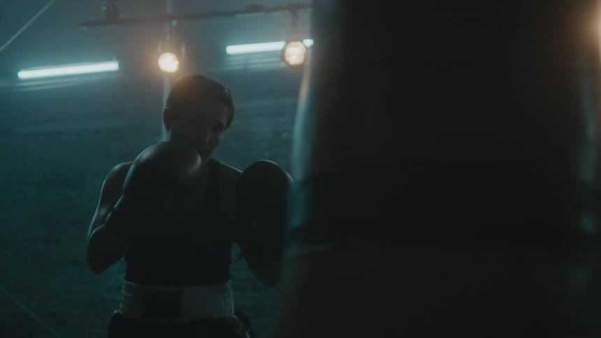 Female boxer with braids wearing a sports bra, fight trunks, and boxing gloves hits punching bag in the gym
