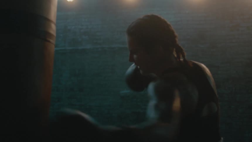 Female boxer with braids wearing a sports bra, fight trunks, and boxing gloves hits punching bag in the gym