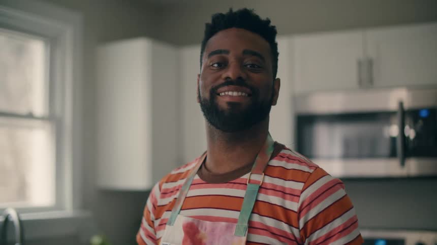 A man is smiling in his kitchen next to a window