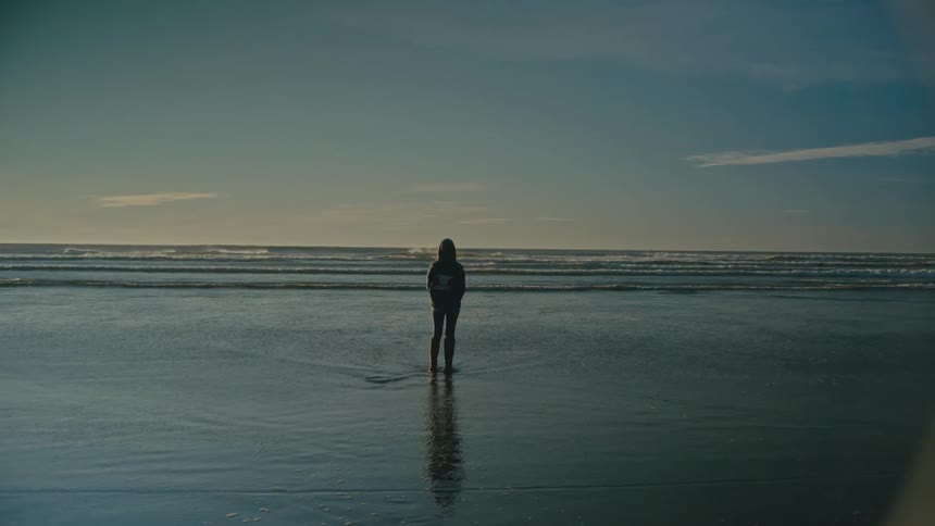 A woman wearing wellington boots stands on a beach while facing the incoming tide