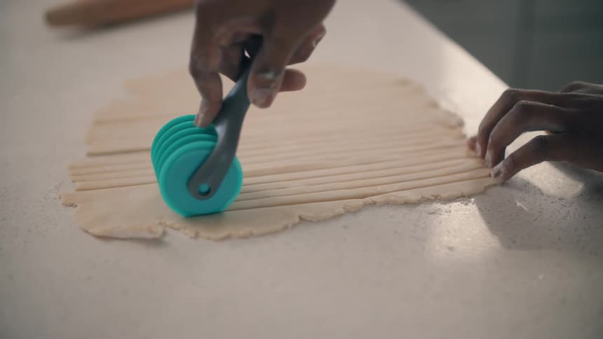 A man is slicing strips of dough on a kitchen counter top with a cutting tool