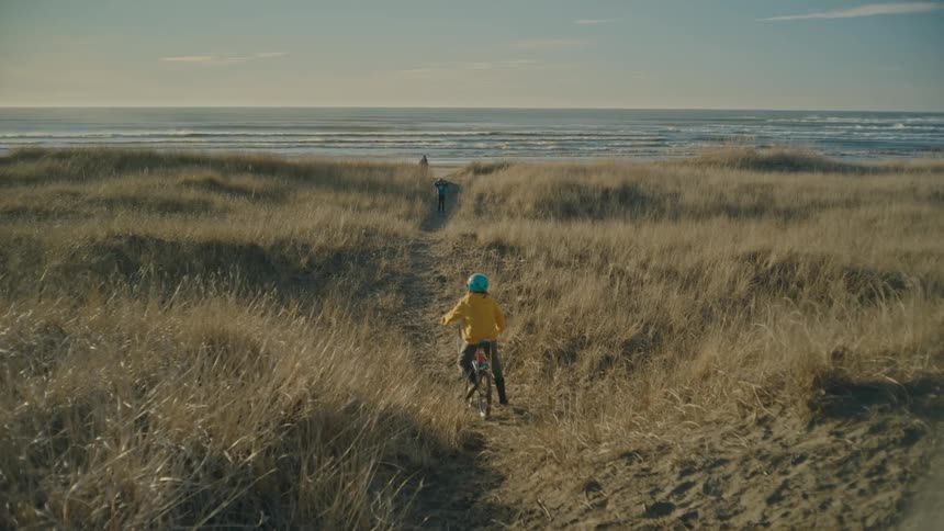 A child is riding a bicycle on a grassy trail towards the ocean